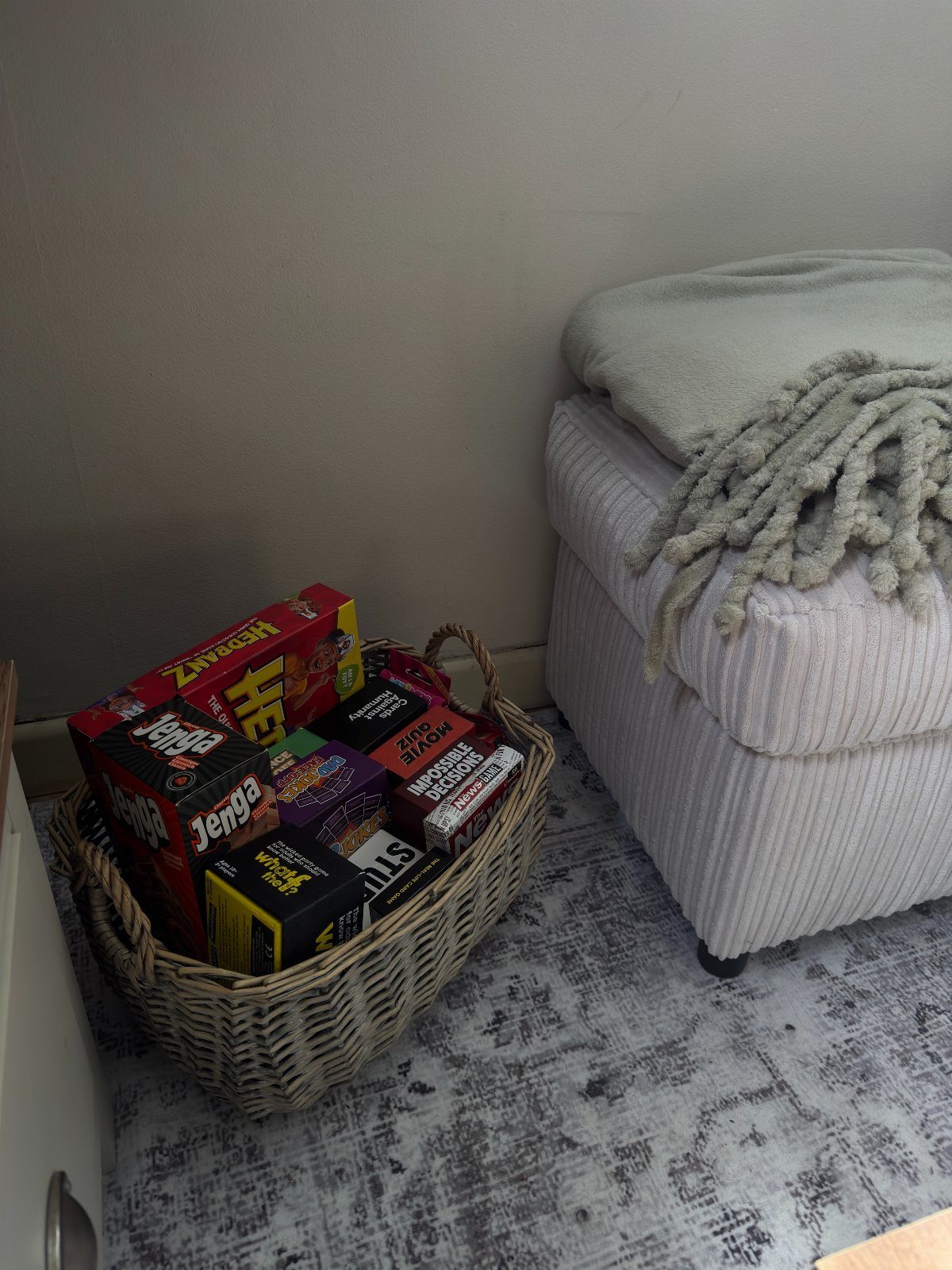 Wicker basket filled with board games sitting on a rug next to an ottoman.