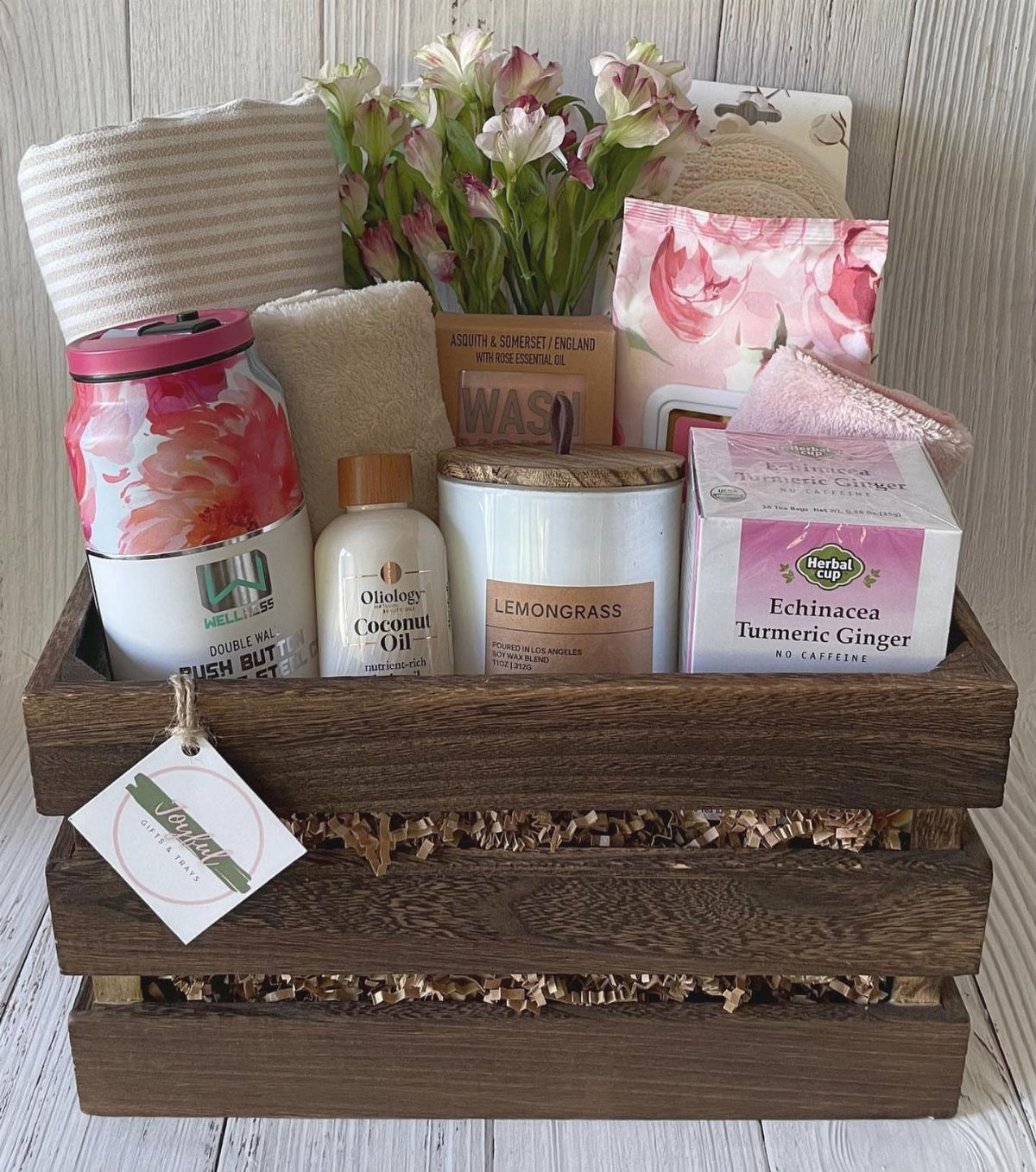 Wooden gift crate containing a candle, herbal tea, coconut oil, and fresh flowers.