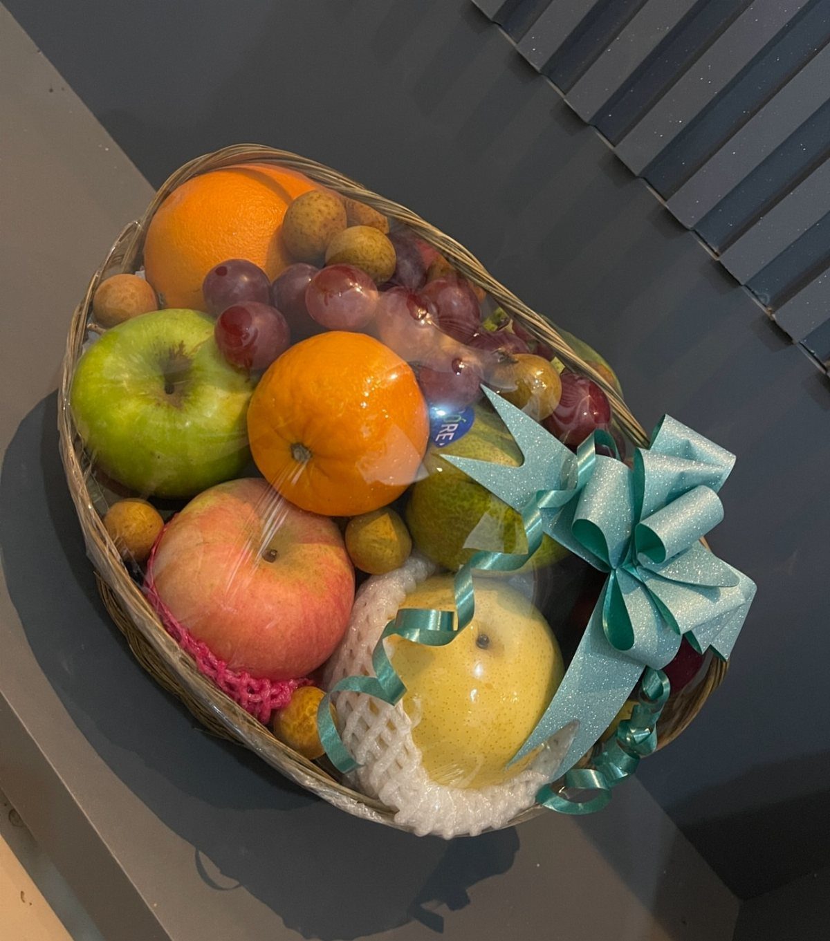 Woven fruit basket with apples, oranges, and grapes tied with a blue ribbon.