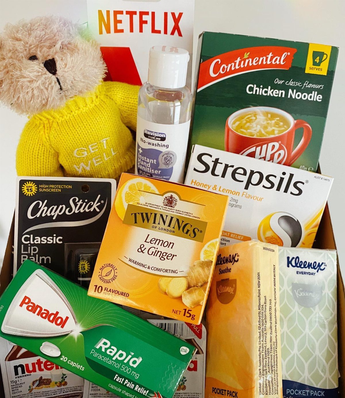Care package box containing a teddy bear, soup, tea, and various health supplies.