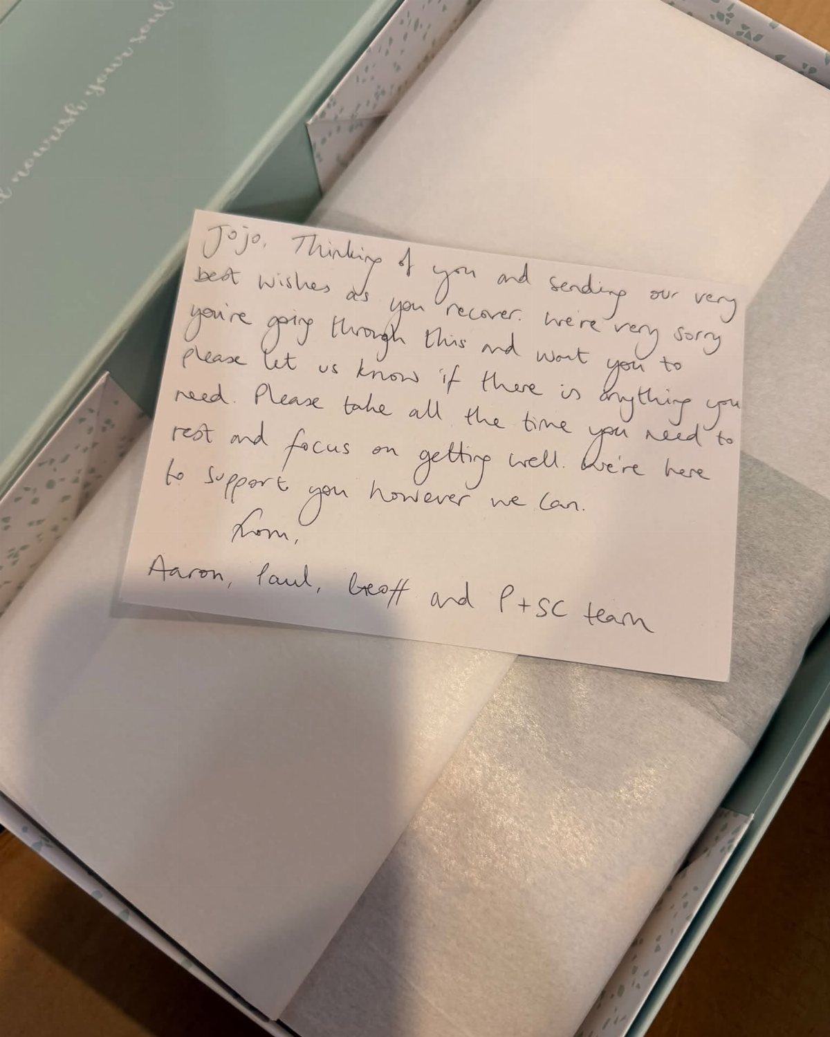 Handwritten get well note resting inside a light blue gift box with tissue paper.