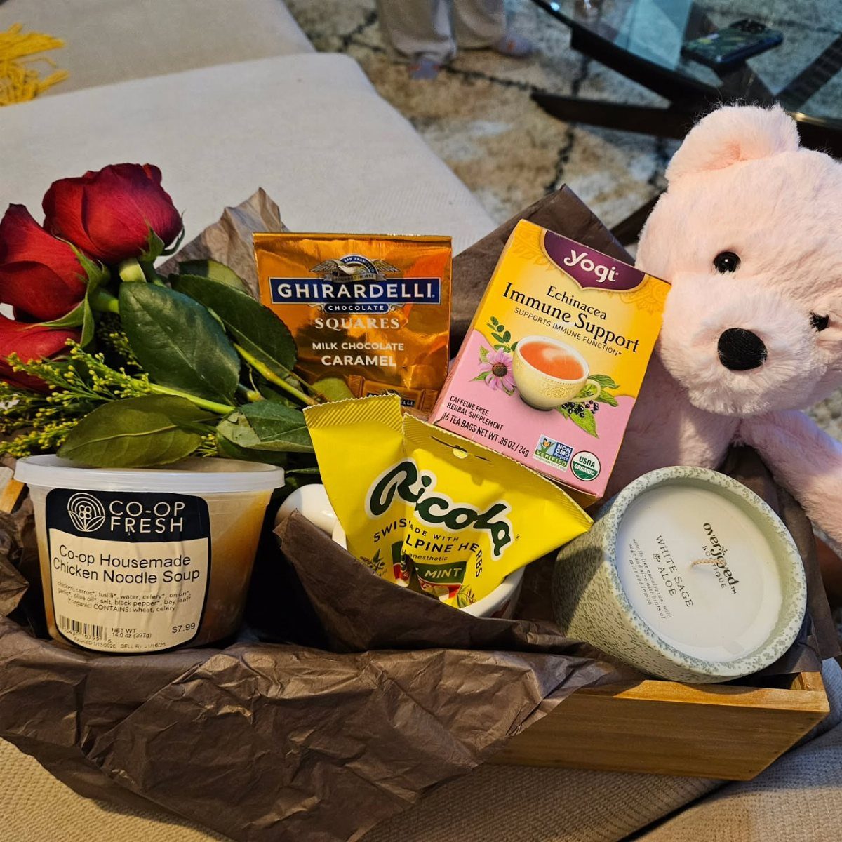 Gift basket with red roses, chicken noodle soup, tea, chocolate, and pink teddy bear.
