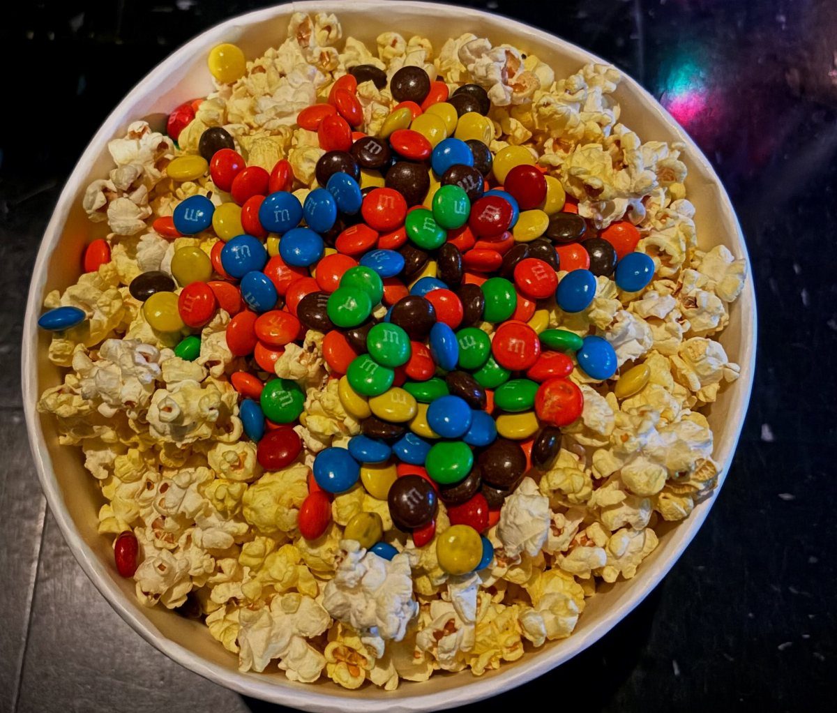 Popcorn and colorful candy-coated chocolates in a white bowl on a dark surface.