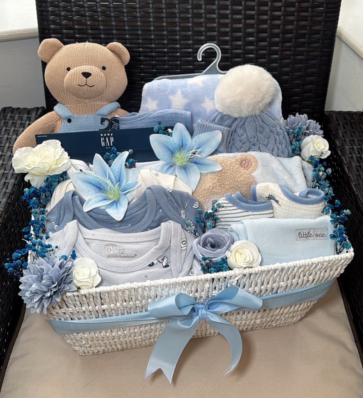 White wicker gift basket filled with blue baby clothes, a teddy bear, and flowers.