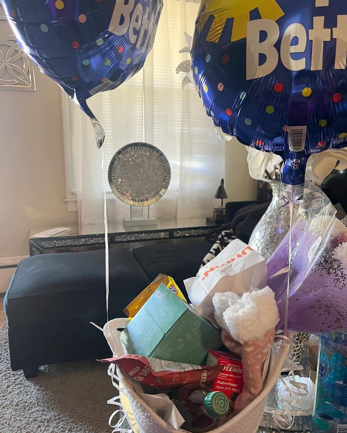 White gift basket with snacks and medicine under two blue personalized balloons.