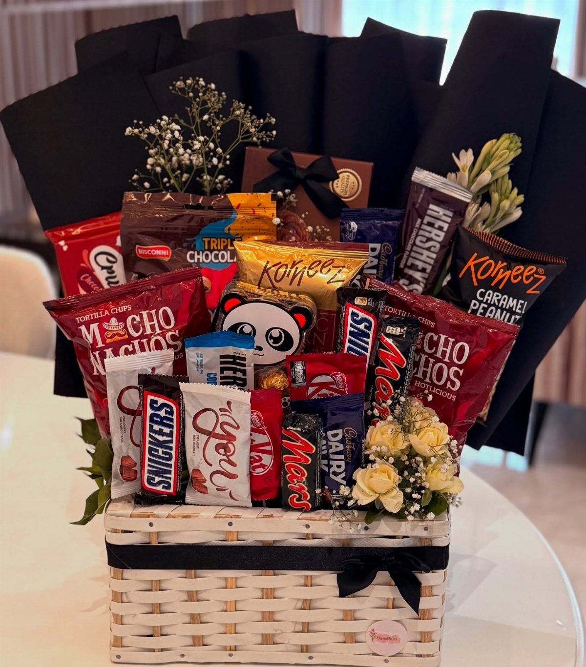 White wicker gift basket filled with assorted chocolates and snacks and white flowers.