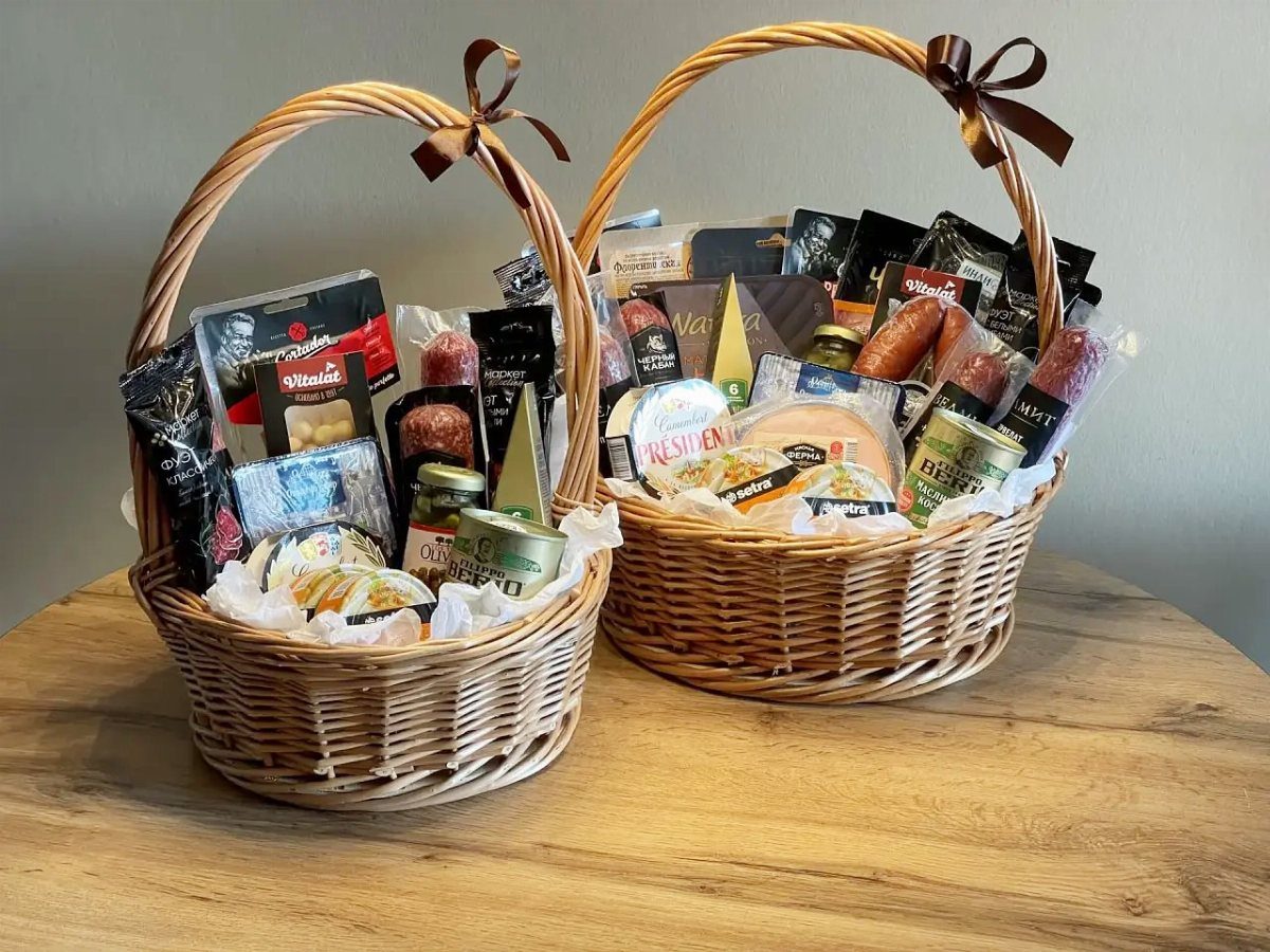 Two wicker gift baskets filled with gourmet cheeses, sausages, and crackers on wood.