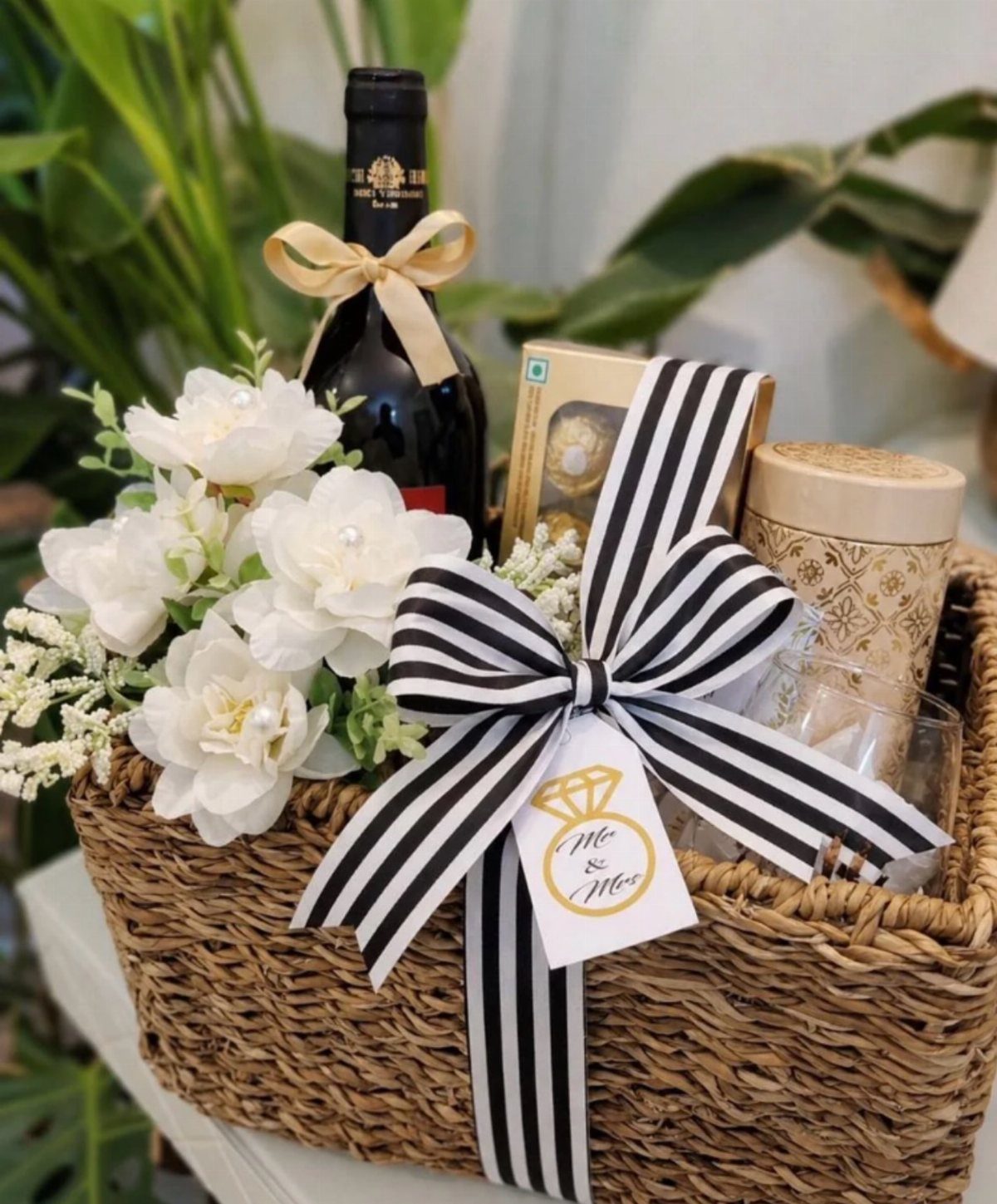Woven gift basket with red wine, white flowers, and a black and white ribbon.