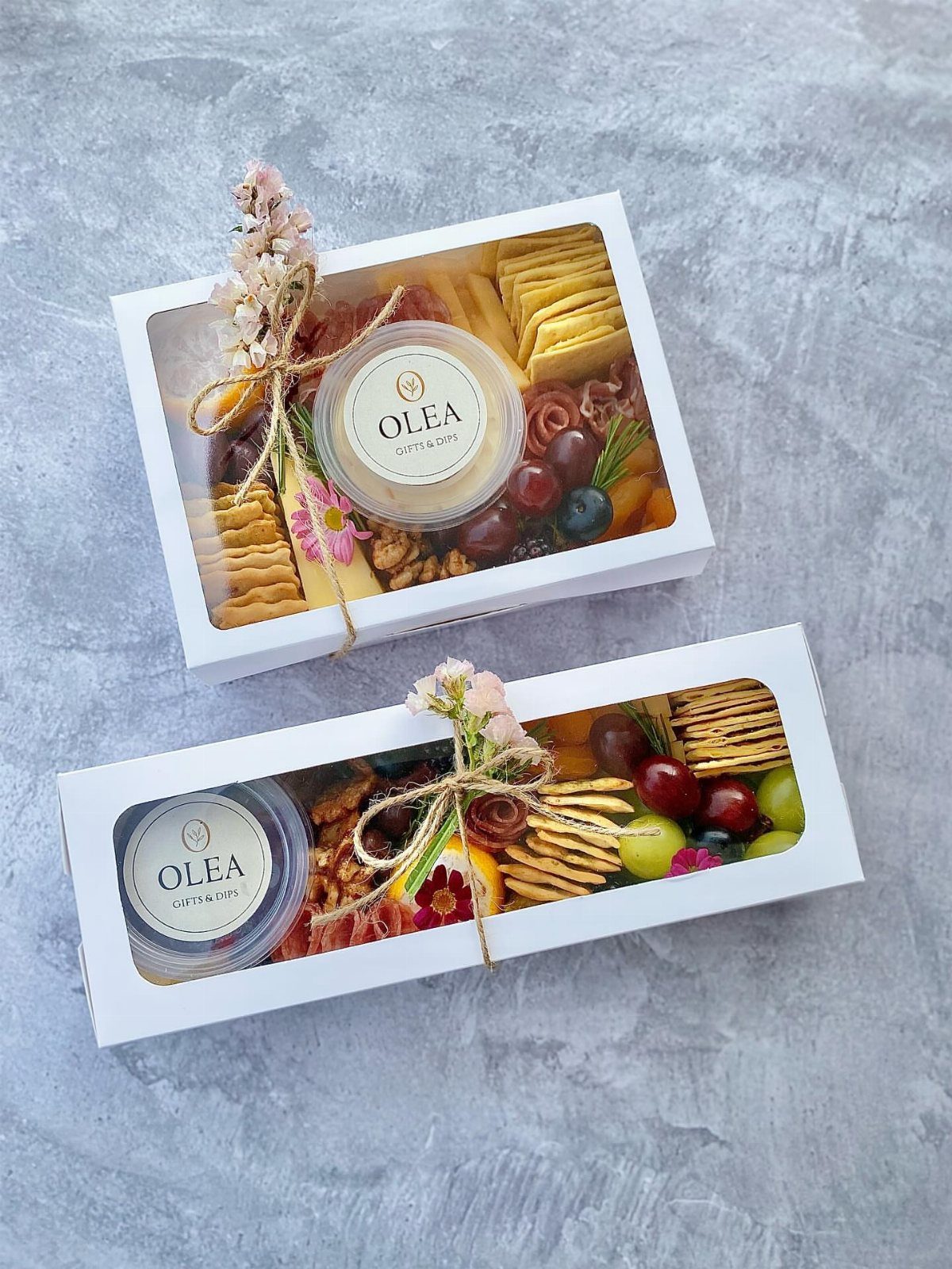 Two white gourmet snack gift boxes filled with crackers, grapes, and dip jars.