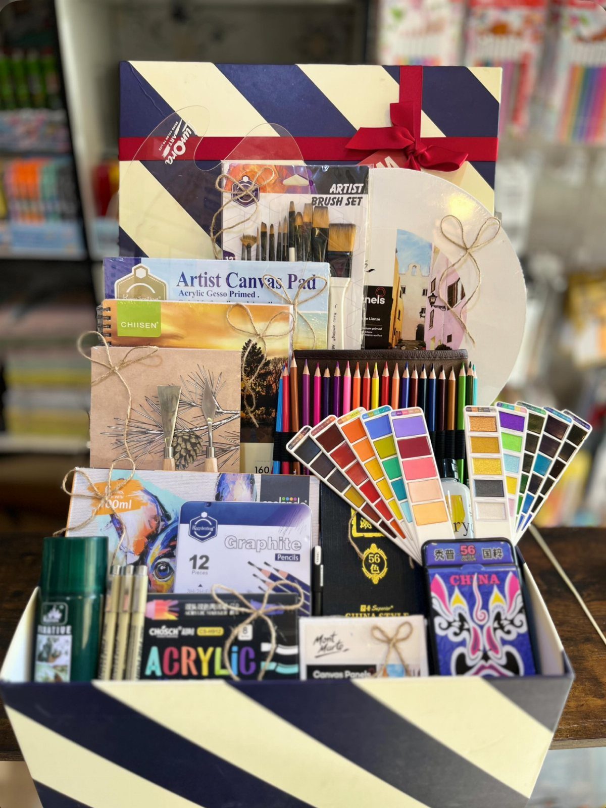 Art supplies gift basket containing brushes, canvas pad, pencils, and paints in striped box.