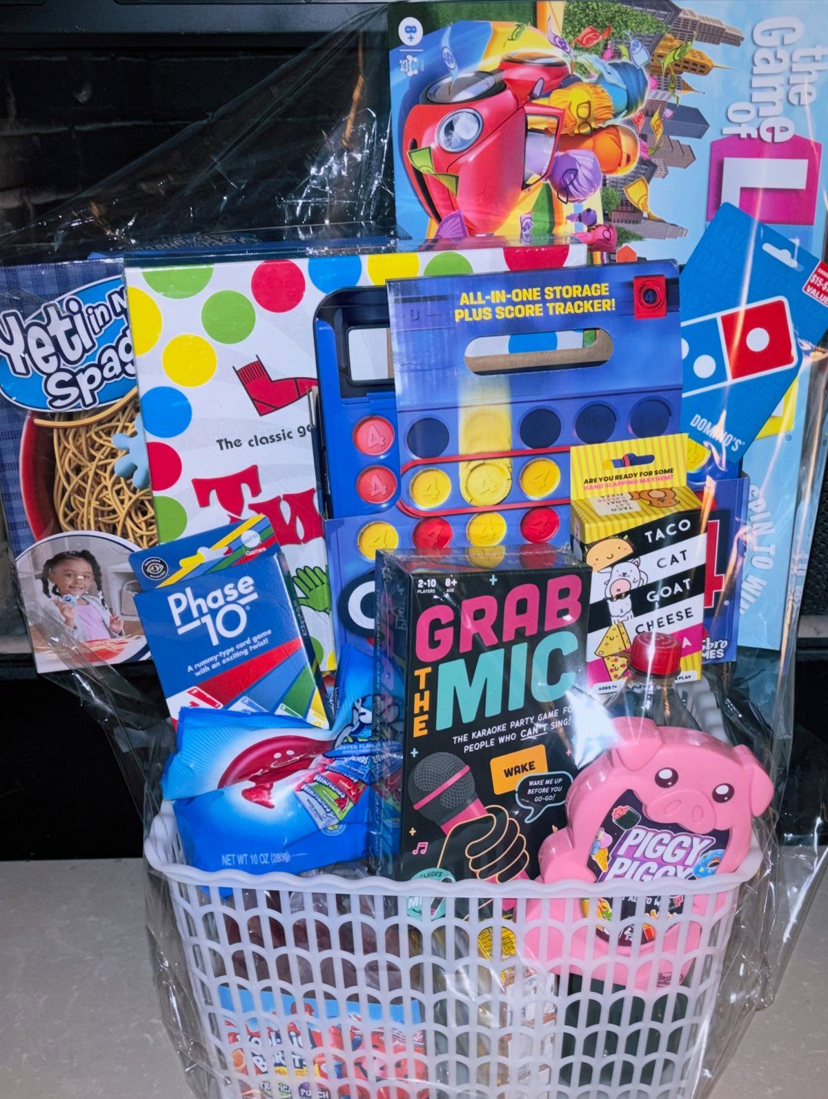 White basket filled with various board games and a pink pig toy wrapped in cellophane