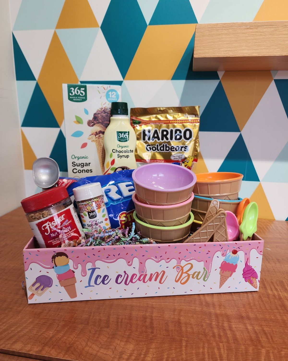 Ice cream bar gift basket with cones, bowls, toppings, and syrup in a tray.