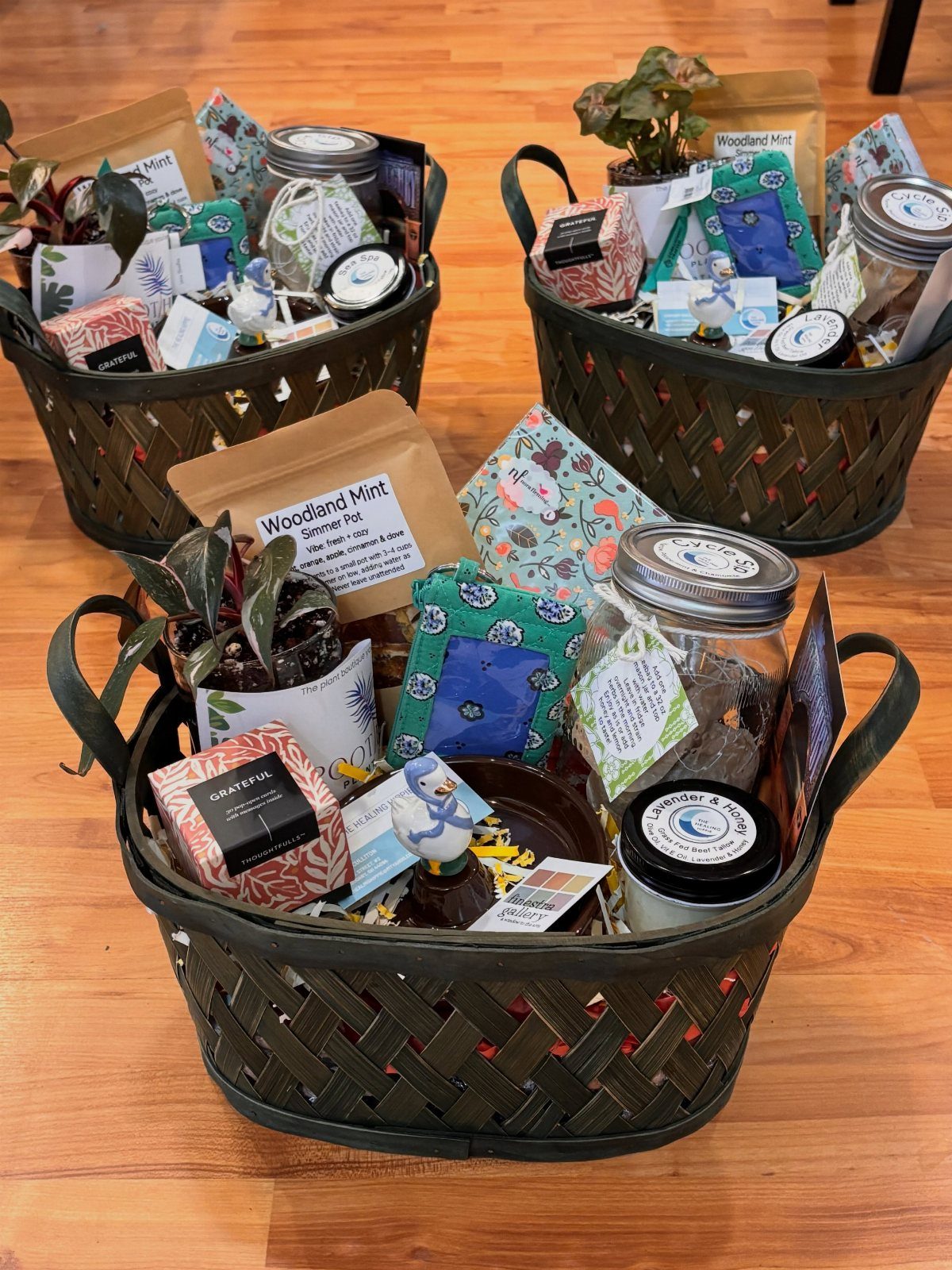 Three woven gift baskets containing plants, jars, and herbal tea on wooden flooring.