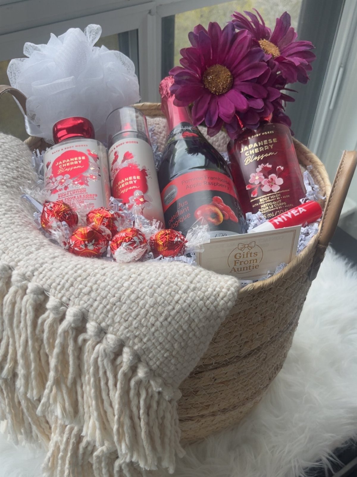 Woven gift basket with spa products, sparkling wine, chocolates, and purple flowers.
