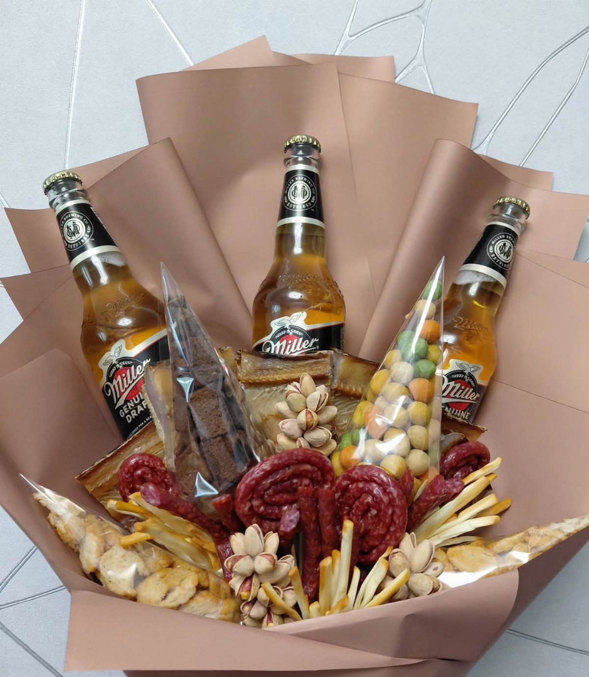 Beer gift arrangement with three Miller bottles, sausages, and snacks in brown paper.