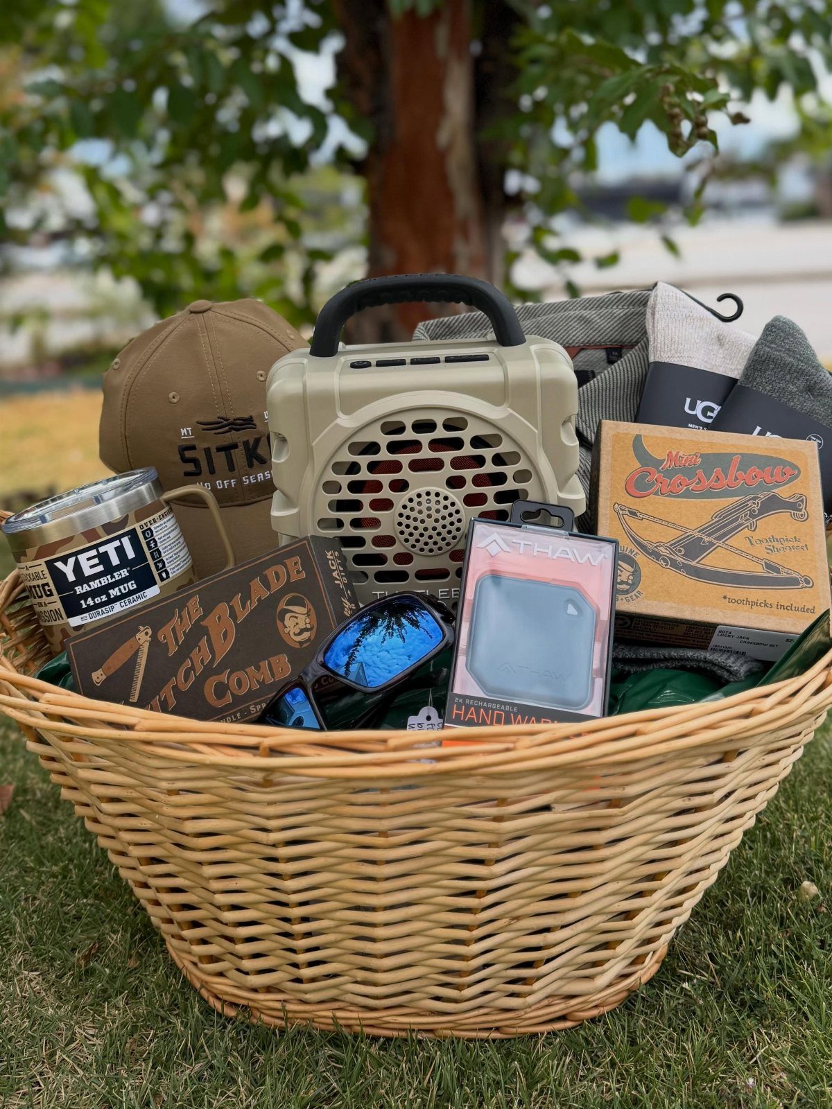 Woven wicker basket filled with outdoor gear, speaker, and cap on green grass.