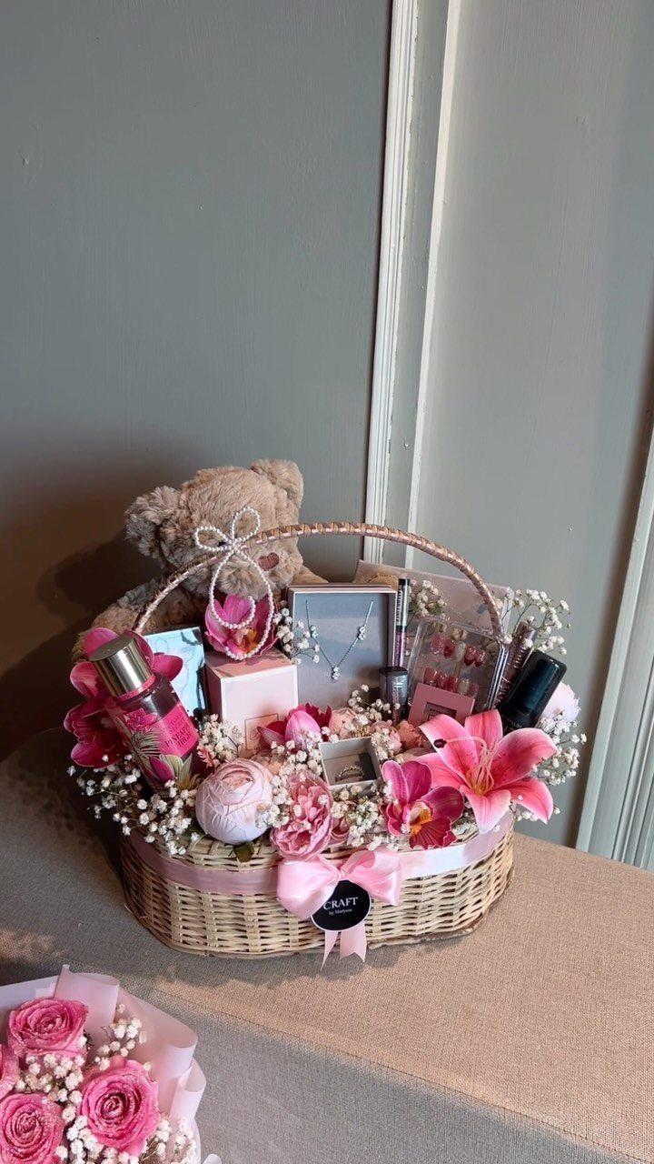 Wicker gift basket with pink flowers, teddy bear, and beauty products tied with ribbon.