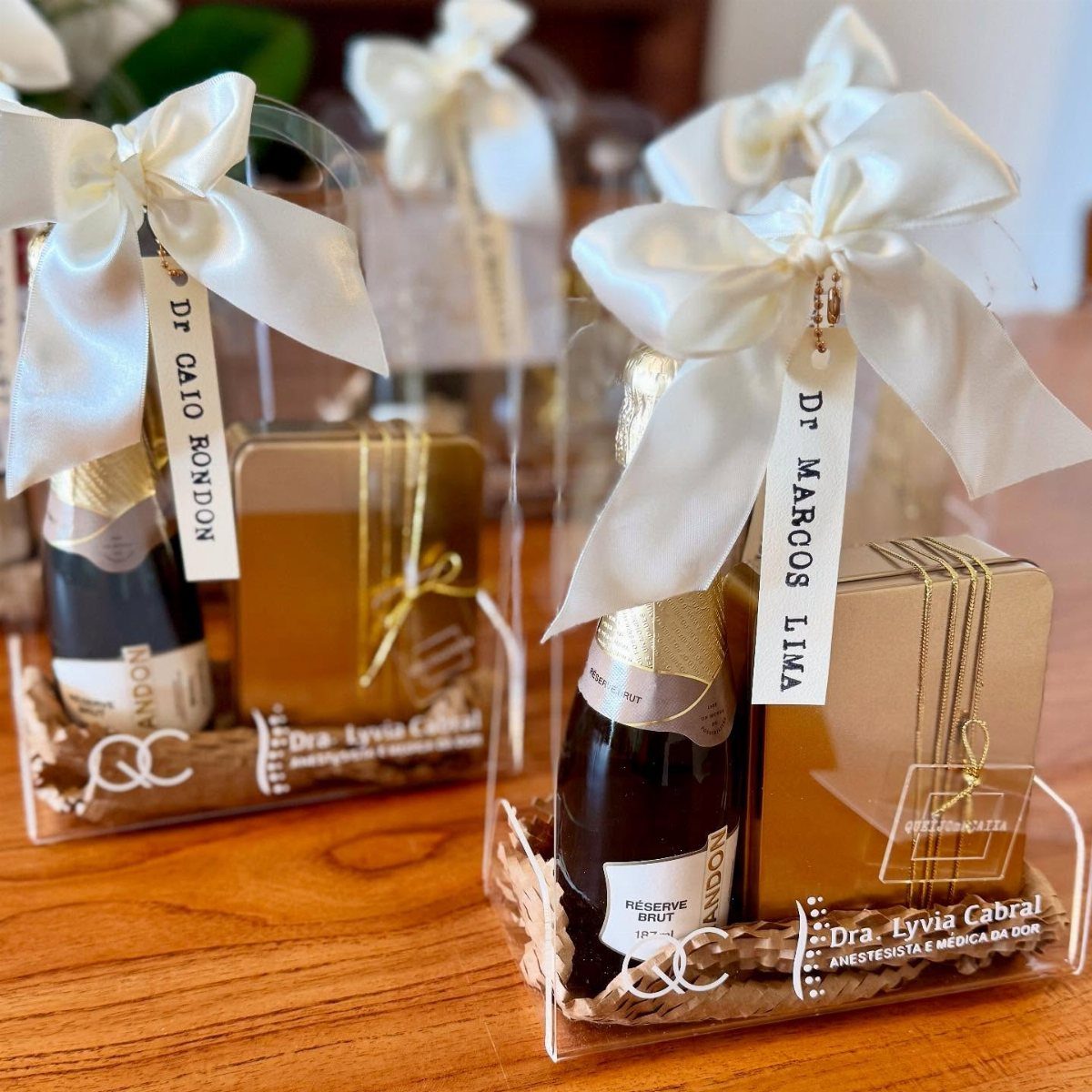 Wine gift baskets in clear acrylic boxes with gold gifts and white bows.