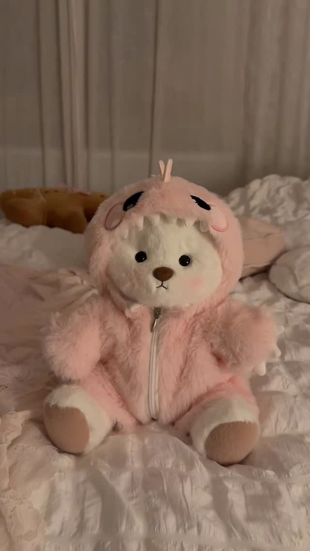 Fluffy white teddy bear in a pink plush hooded jumpsuit on white bedding.