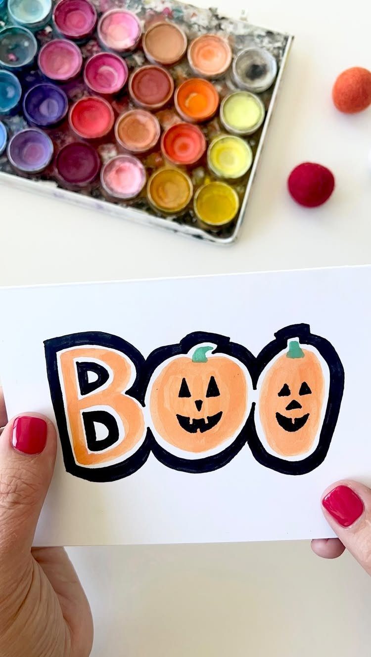 Hand-drawn Halloween card with BOO and pumpkins held over watercolor paints.