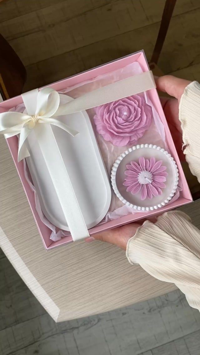 Pink gift box containing floral candles and a white ribboned object held by hands.