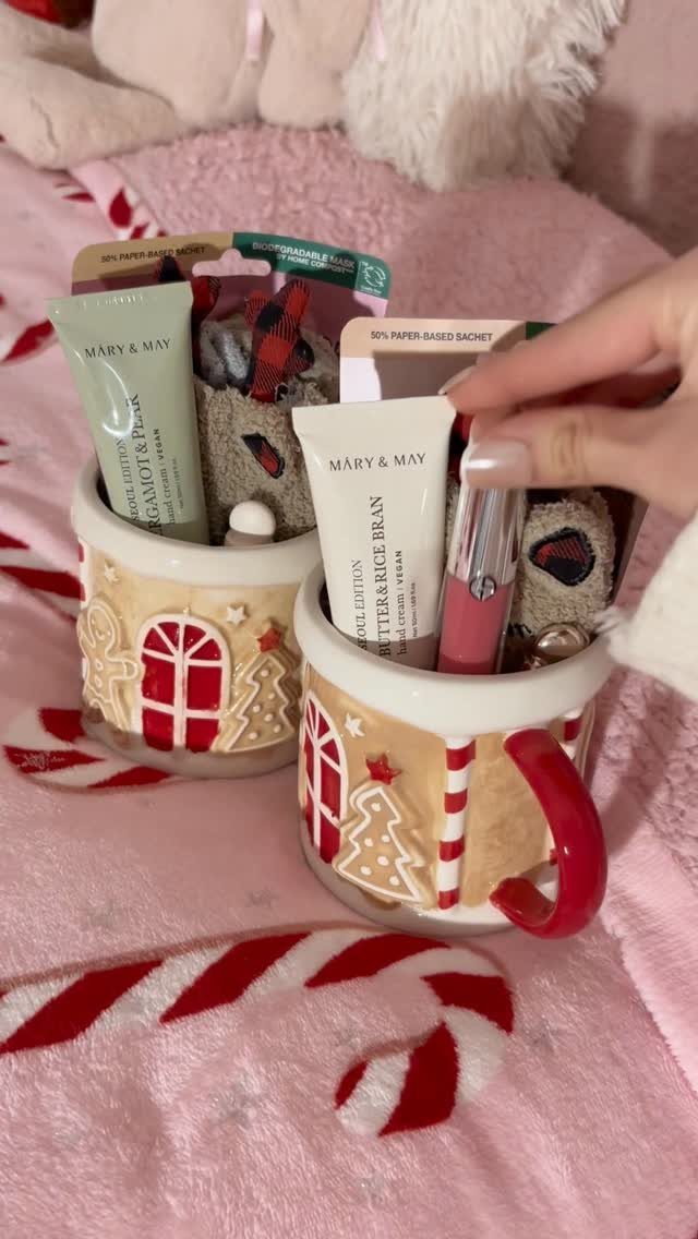 Two gingerbread-themed ceramic mugs filled with skincare products on a pink fuzzy blanket.