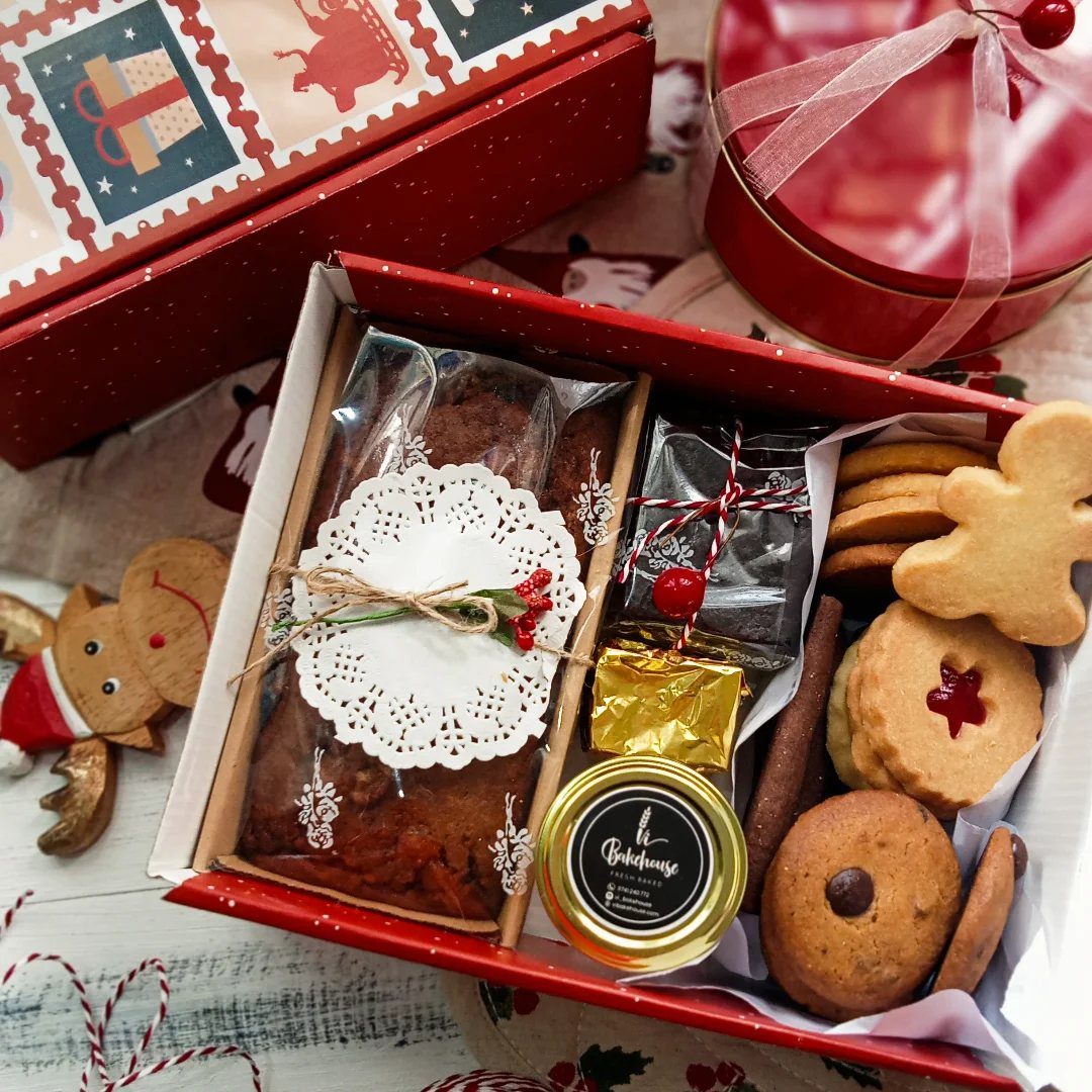 Red gift box containing fruitcake, jam, and assorted Christmas cookies on white wood.
