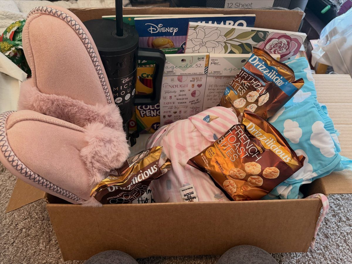 Care package in a cardboard box containing pink slippers, snacks, and a travel mug.