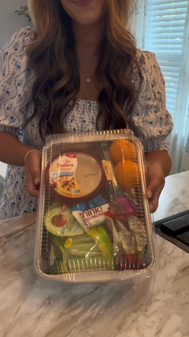 Clear plastic container filled with hummus, oranges, and healthy snacks held by person.