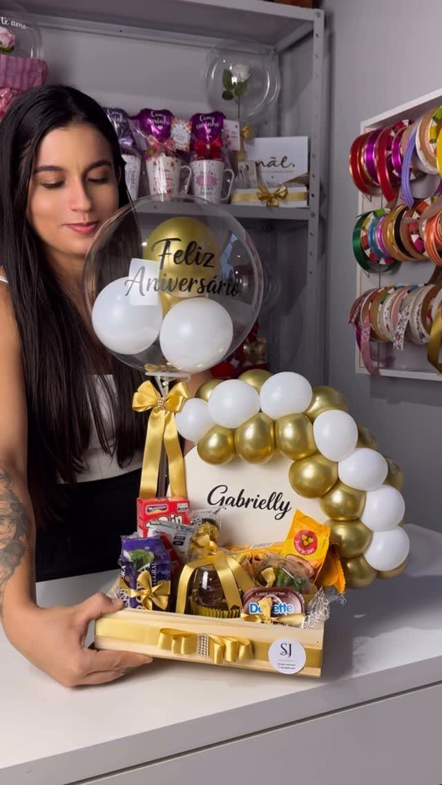 Woman holding a gift basket with gold and white balloons and snacks.
