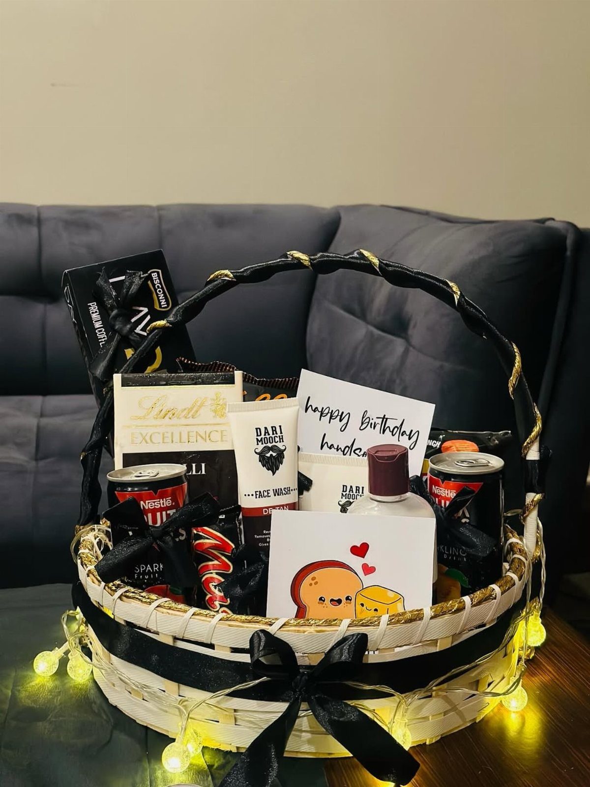 White wicker gift basket with chocolates and skincare, decorated with black ribbons and fairy lights.