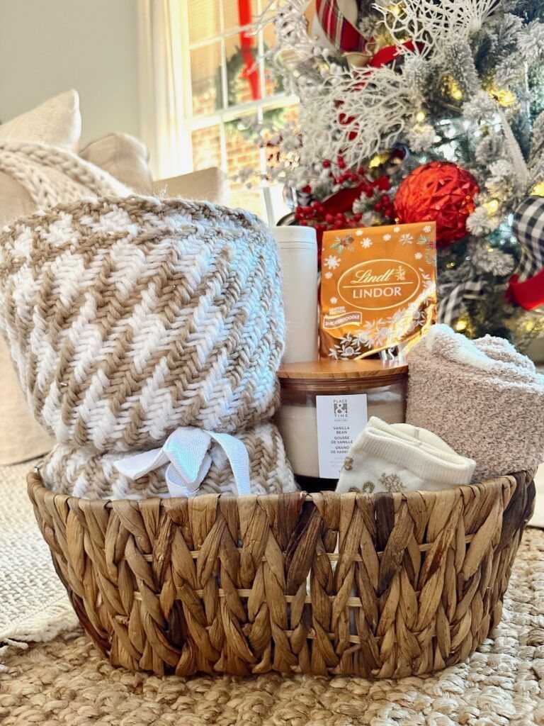 Woven gift basket with a patterned blanket, chocolates, and a candle near a Christmas tree.