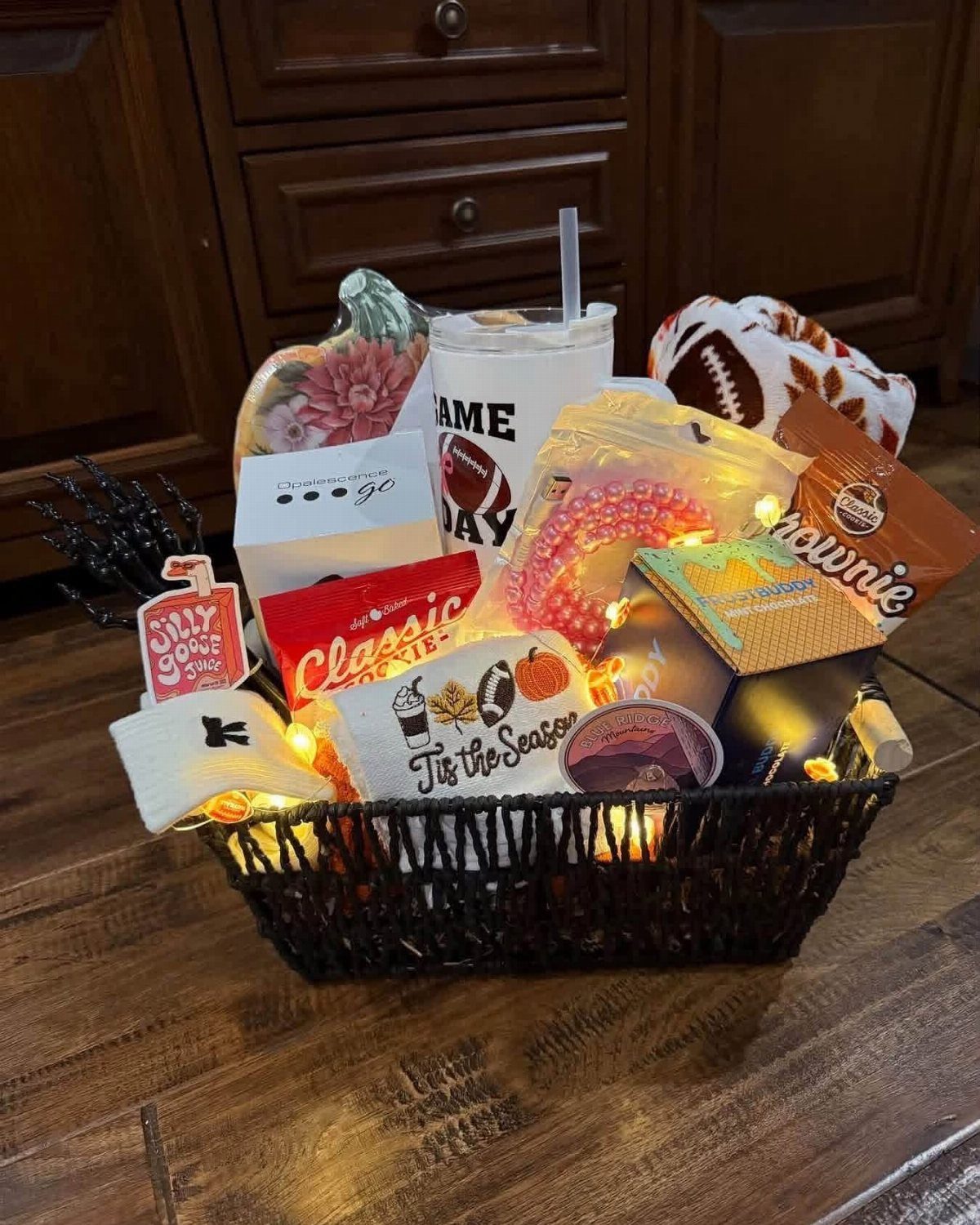 Black woven gift basket filled with snacks, a tumbler, and warm fairy lights.