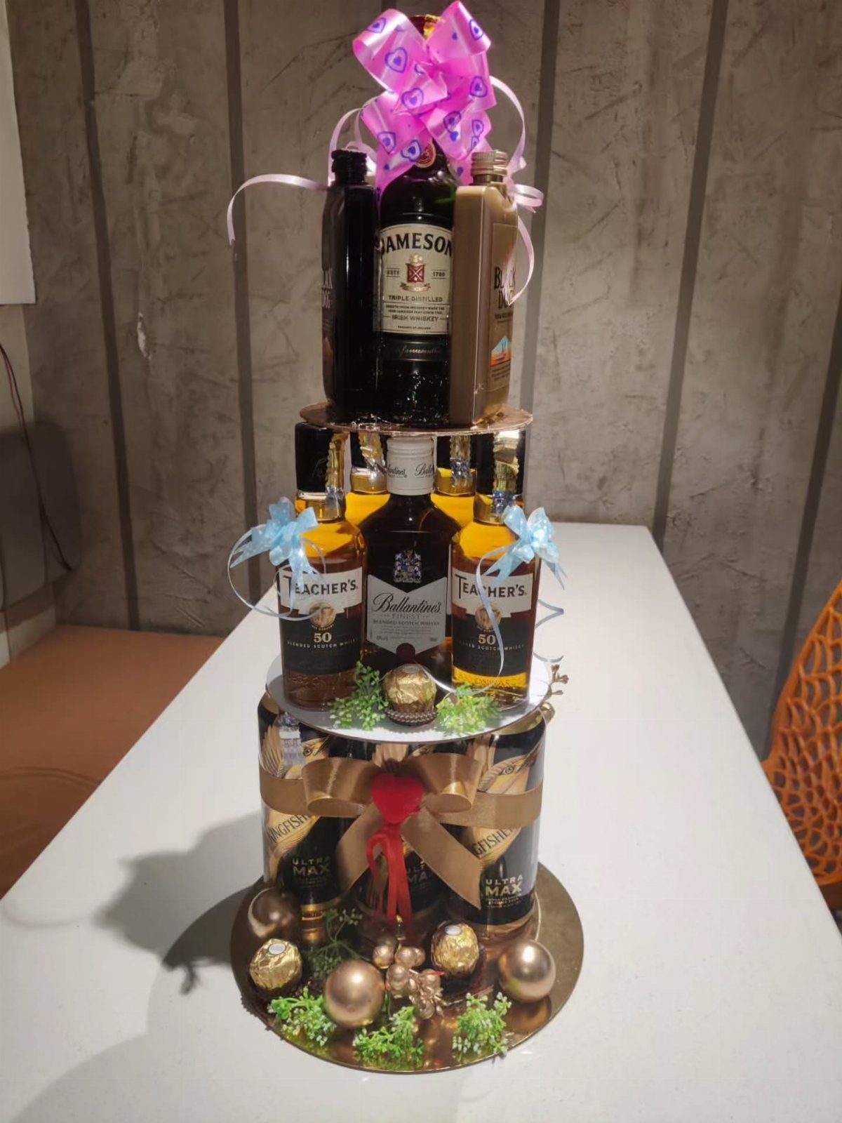 Tiered liquor bottle tower decorated with ribbons and gold ornaments on a white table.