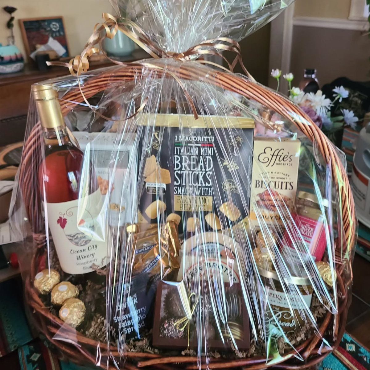 Wicker gift basket with rosé wine, gourmet chocolates, and savory snacks wrapped in cellophane.