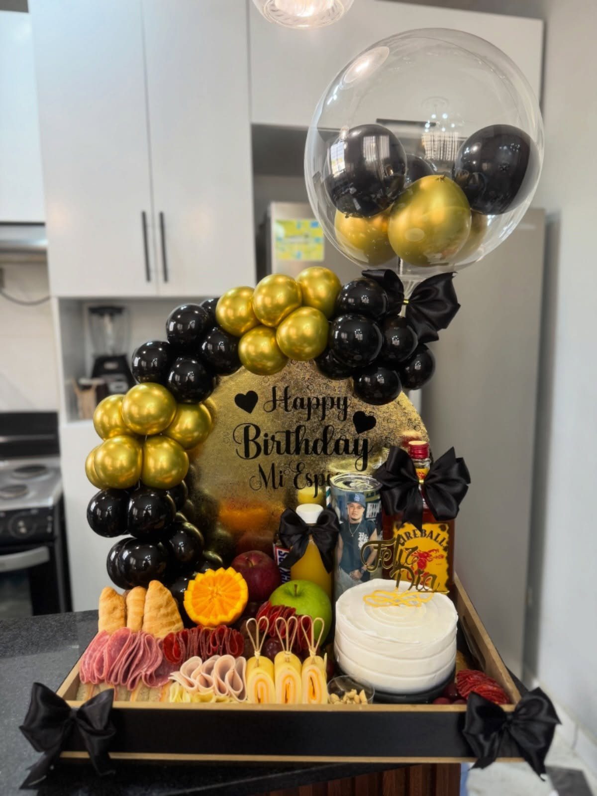 Birthday gift tray with charcuterie, cake, and black and gold balloon decorations.