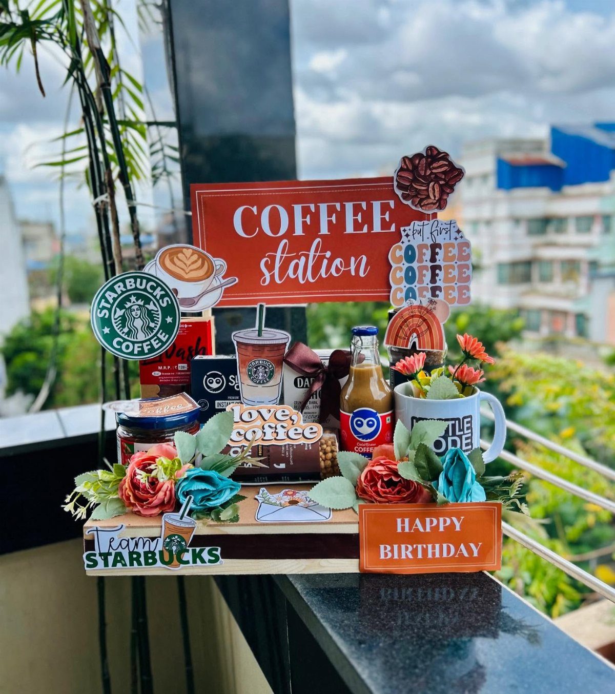 Coffee gift basket with Starbucks items, a white mug, and birthday signs on a balcony.