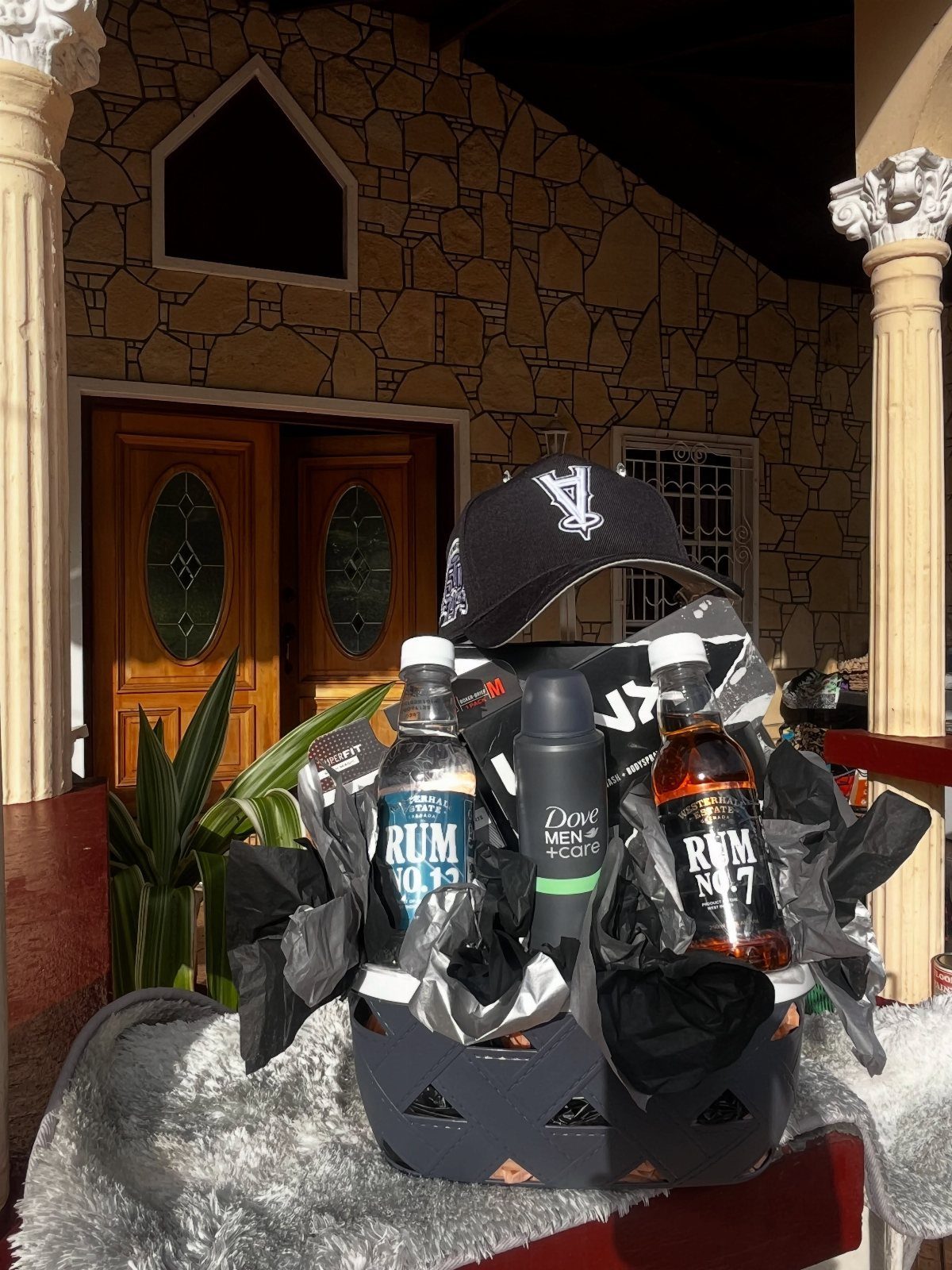 Gift basket with rum bottles, deodorant, and a black cap on a porch.