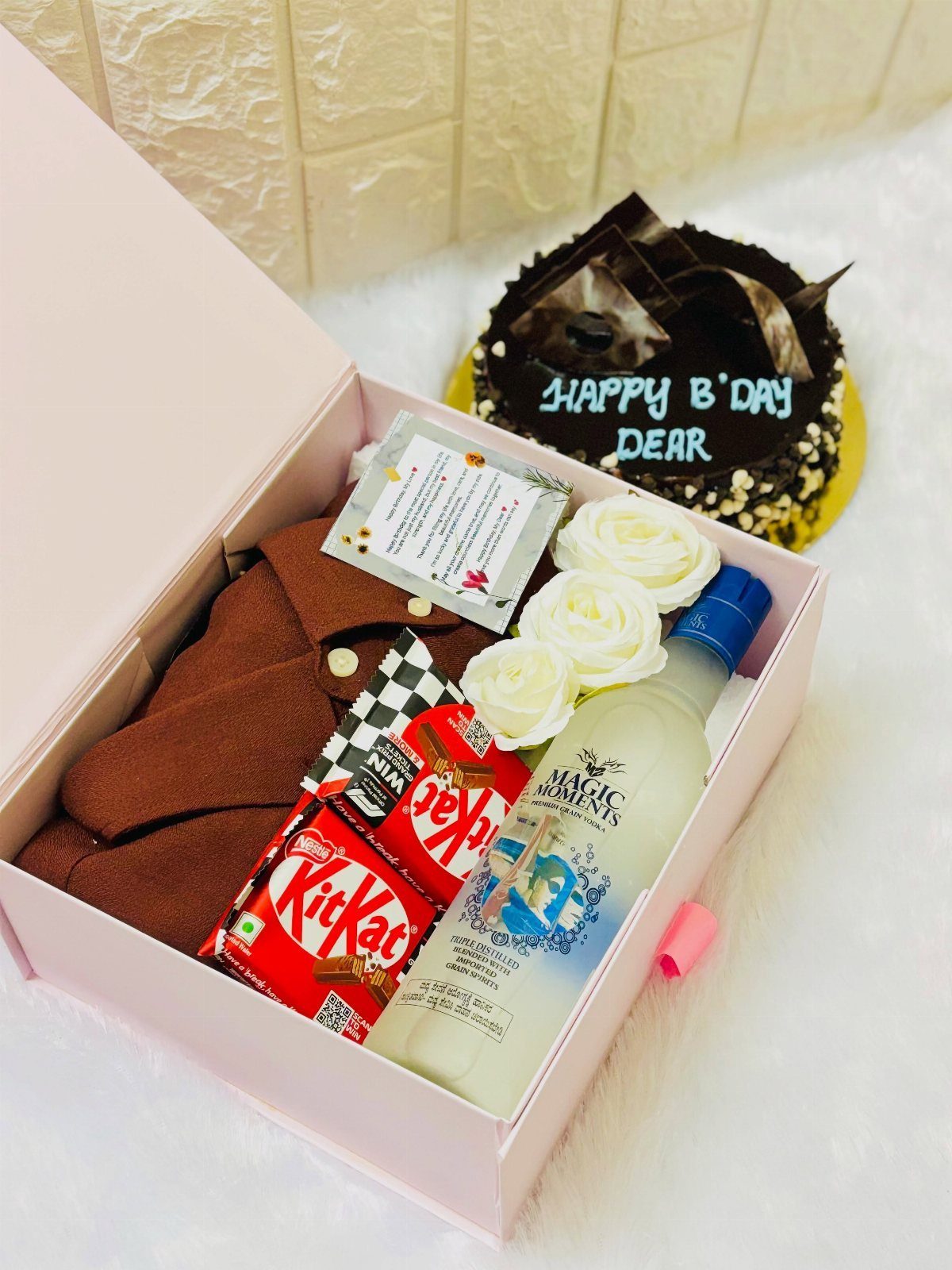 Pink gift box with brown shirt, vodka, chocolates, and roses beside a birthday cake