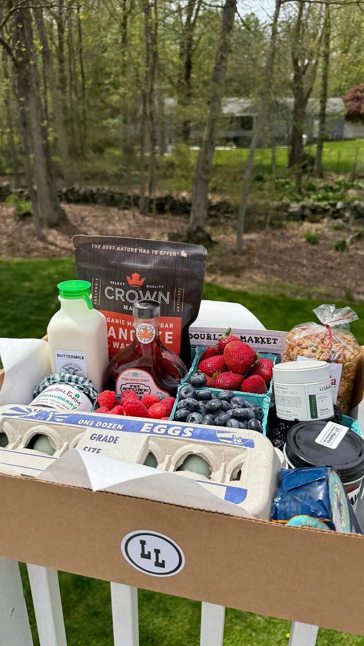 Gift basket with fresh berries, eggs, maple syrup, and milk outdoors in nature.