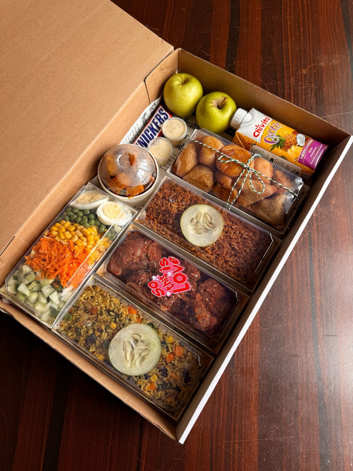 Cardboard gift box filled with assorted meals, fruits, and snacks on wooden table