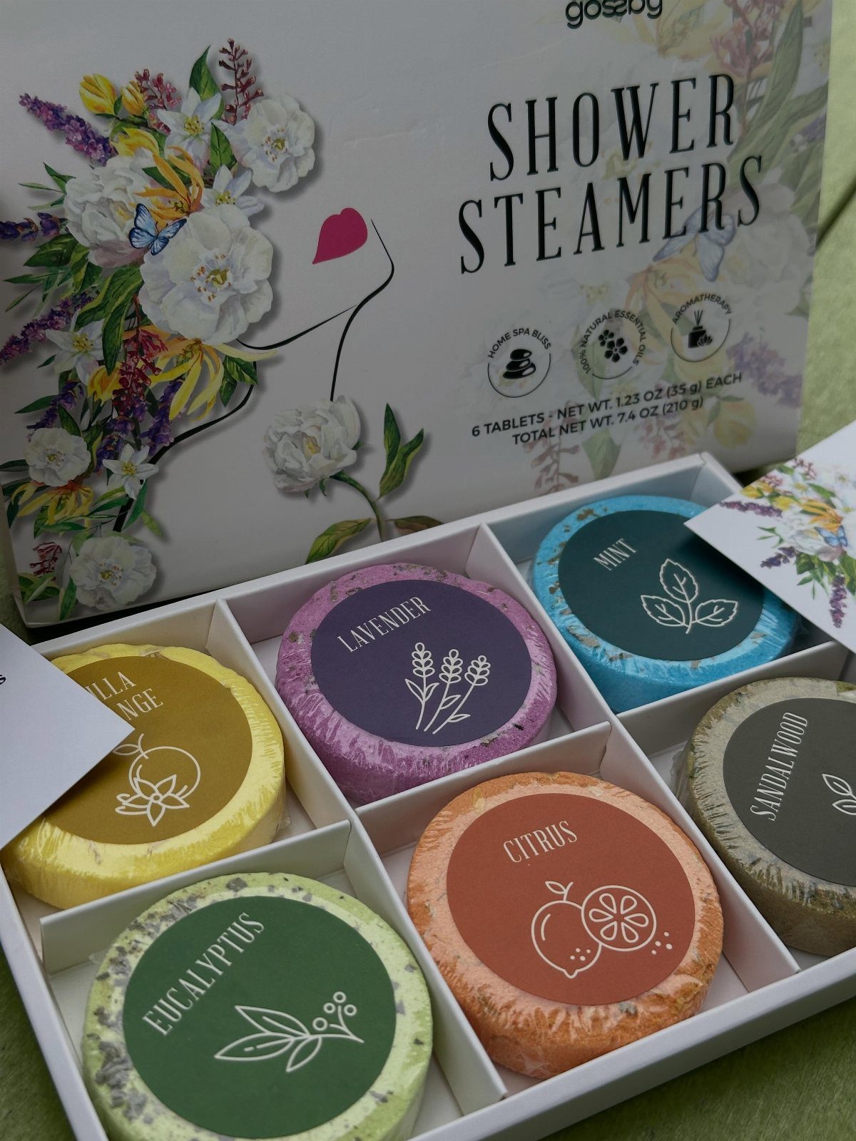 Colorful round shower steamer tablets in a white floral gift box.