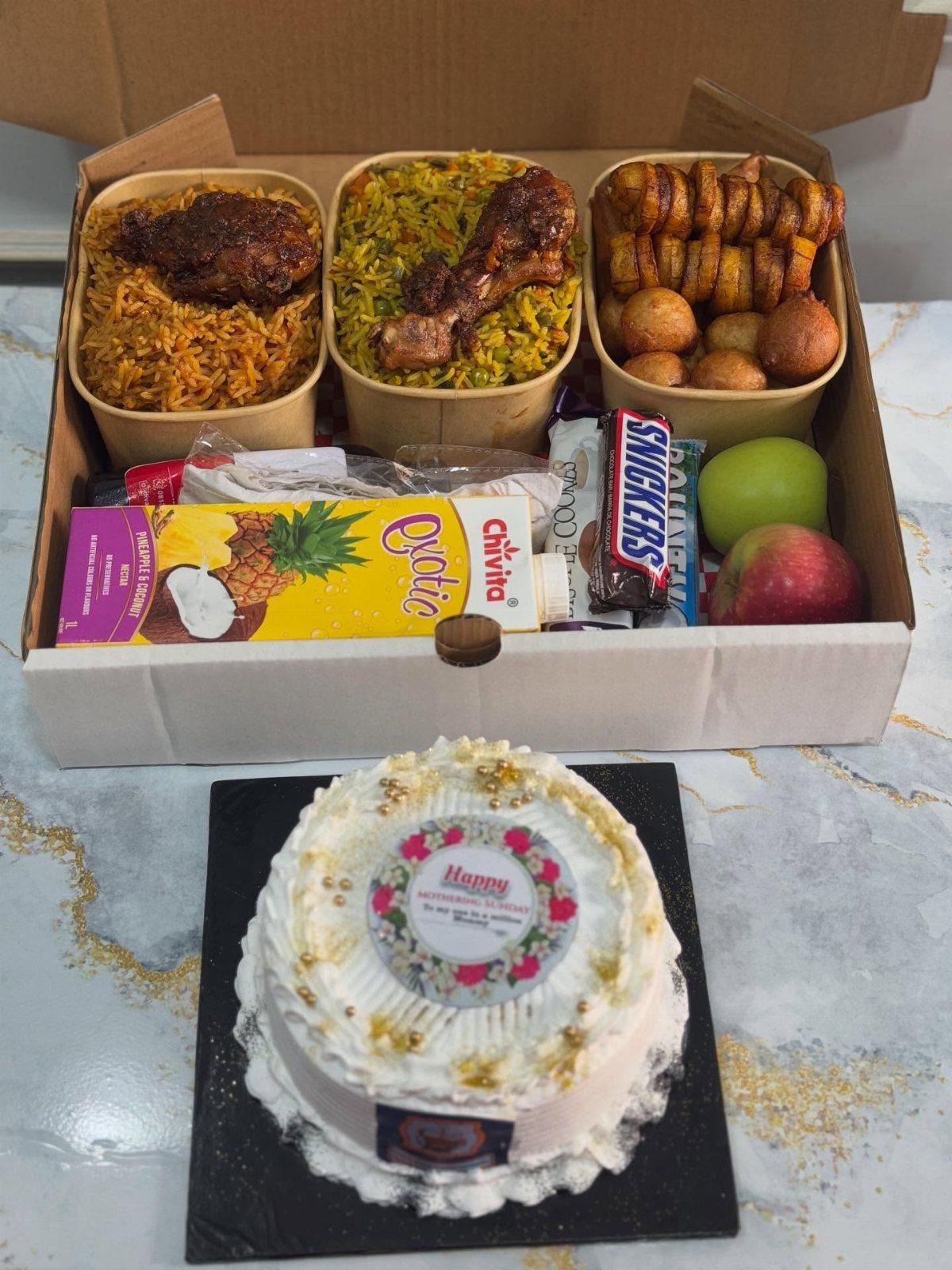 Gift box with savory meals, snacks, and fruit next to a decorated white cake