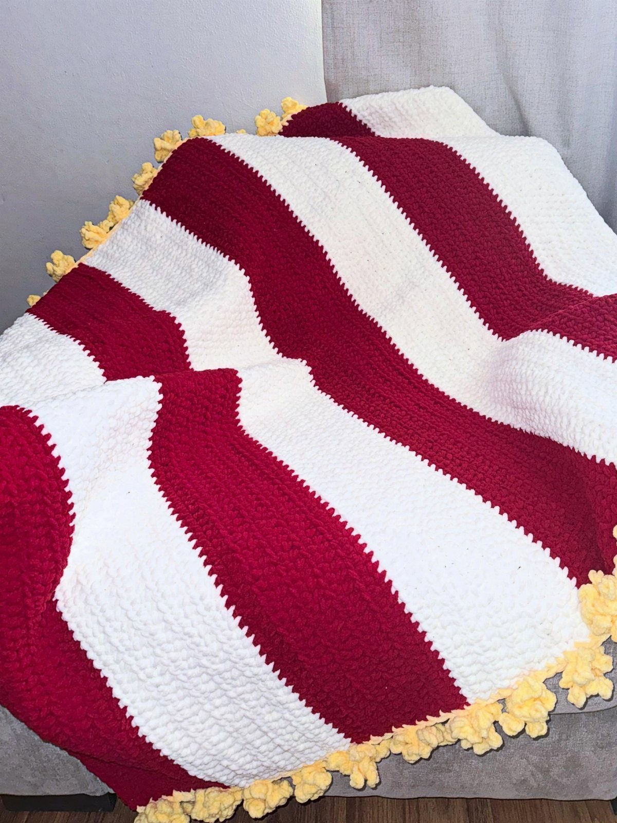 Red and white striped crochet blanket with yellow pom-pom trim on grey fabric.