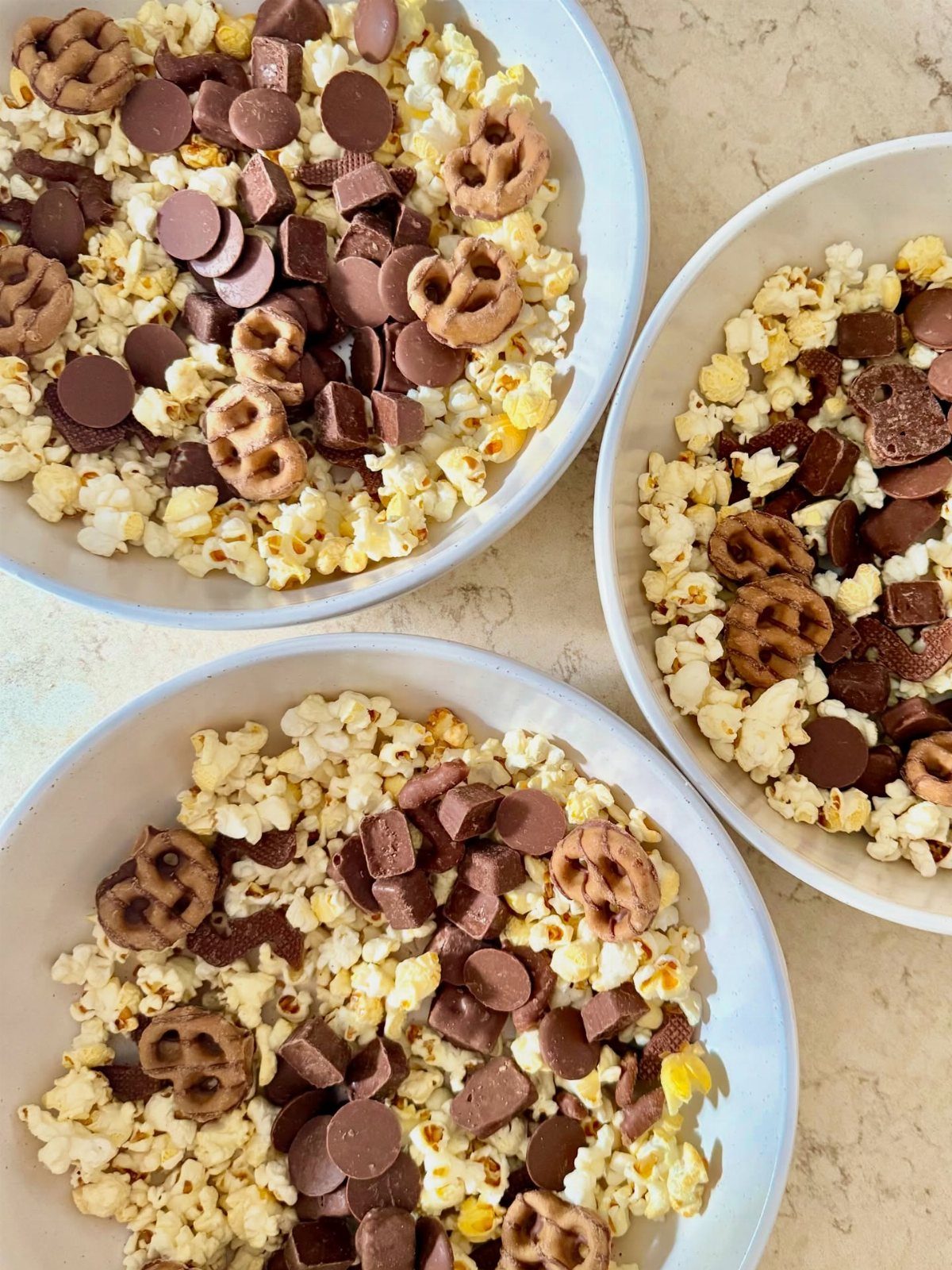 Three white bowls filled with popcorn, chocolate pieces, and chocolate-covered pretzels.