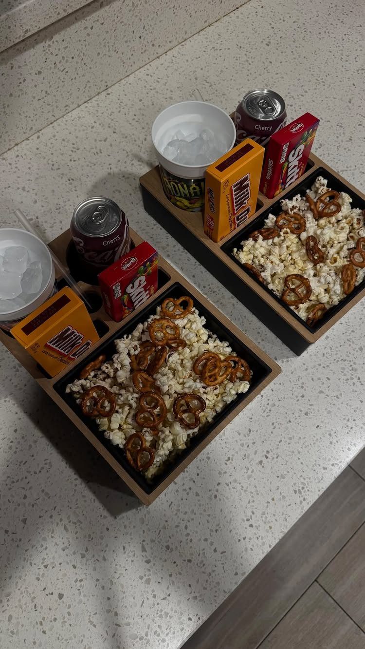 Two wooden snack trays filled with popcorn, pretzels, soda, and candy on a counter.
