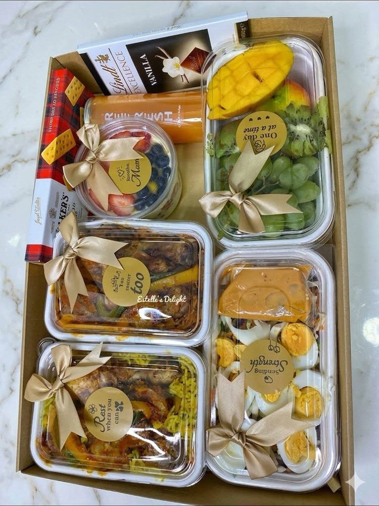 Gift box containing prepared meals, fresh fruit, and care products with gold ribbons.