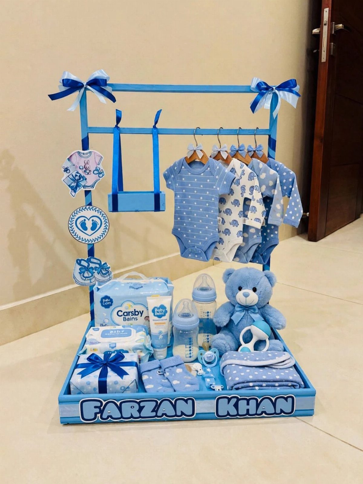 Blue baby gift stand with hanging onesies, teddy bear, and newborn essentials.
