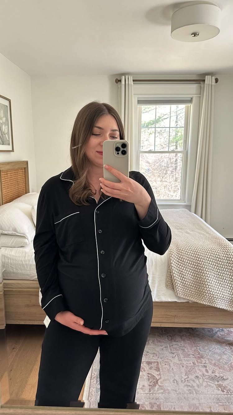 Pregnant woman wearing black button-down pajamas with white piping in a bedroom mirror selfie
