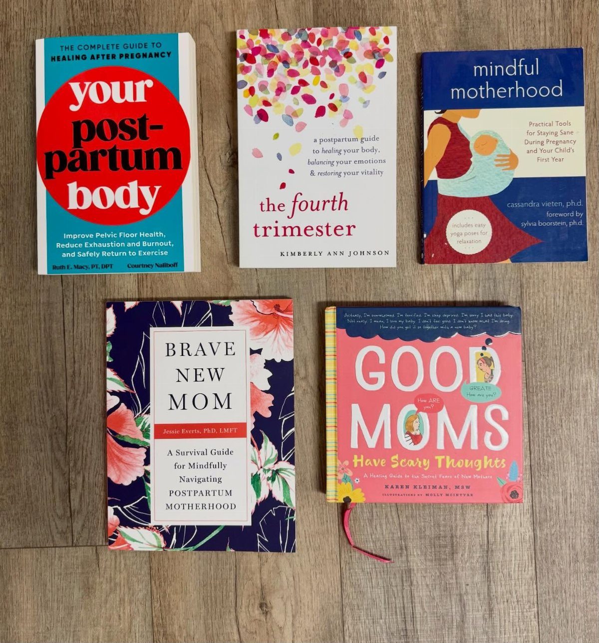 Five postpartum recovery and motherhood books arranged on a light wooden tabletop.